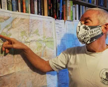 Man wearing a 'coast to coast, scotland' facemask pointing at a map