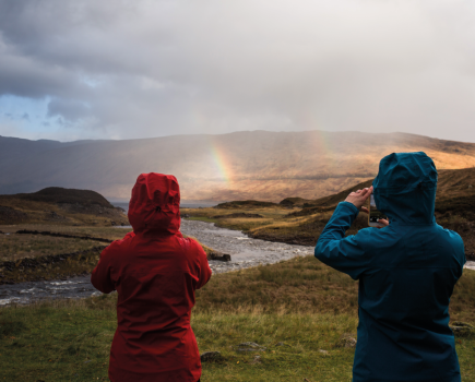 Two people in lightweight waterproofs holding their camera phones and taking pictures of the rainbow in front of them.