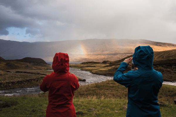 Two people in lightweight waterproofs holding their camera phones and taking pictures of the rainbow in front of them.