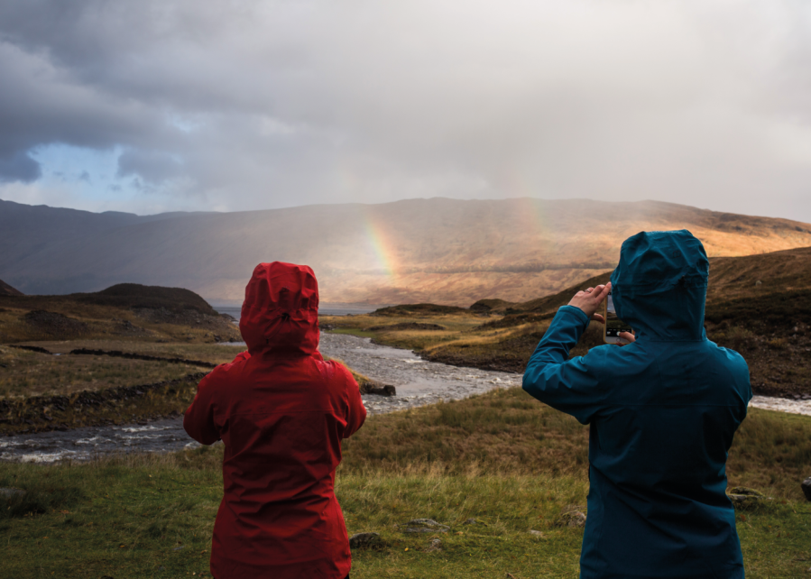 Two people in lightweight waterproofs holding their camera phones and taking pictures of the rainbow in front of them.