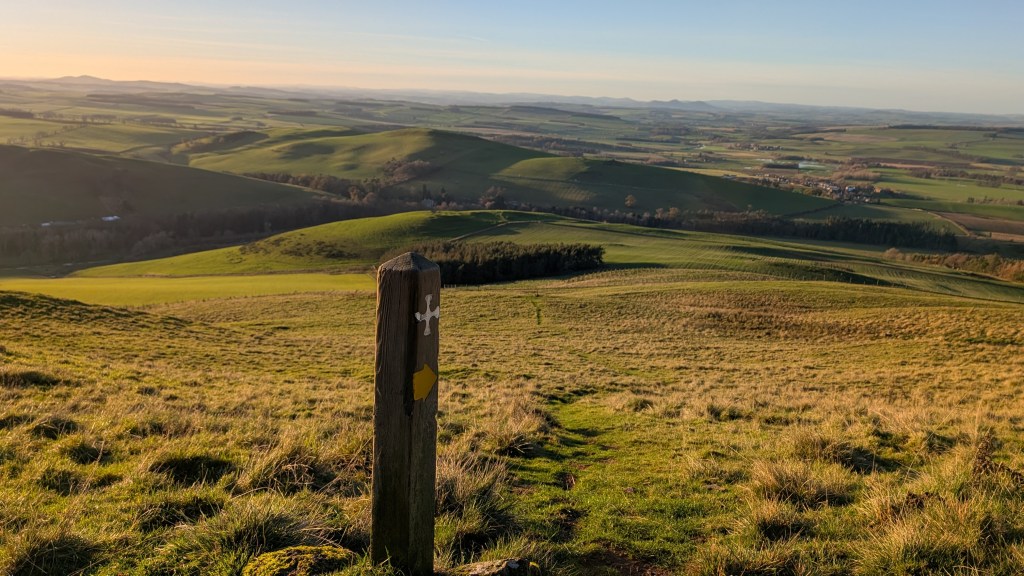 St Cuthbert's Way waymarker and a path leading downhill in the Northern Cheviots looking back towards the village Morebattle