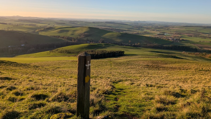 St Cuthbert's Way waymarker and a path leading downhill in the Northern Cheviots looking back towards the village Morebattle
