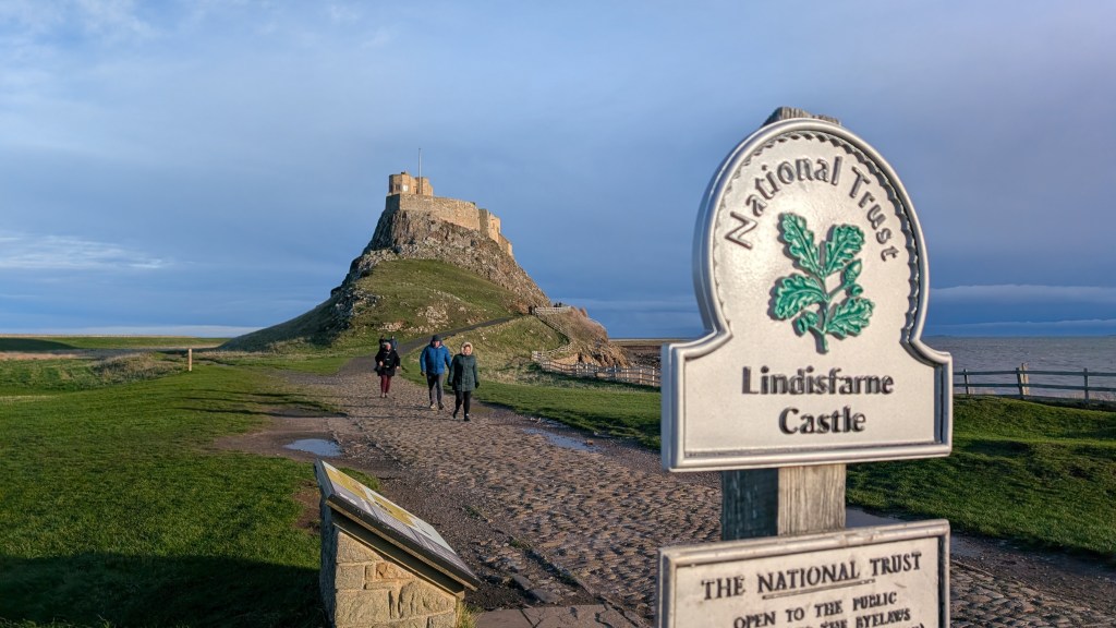 A sign for Lindisfarne Castle with the castle and walkers in the background