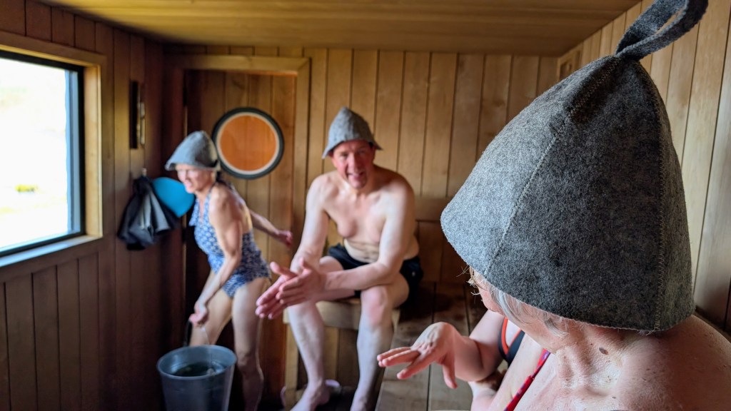 Sauna goers in sauna hats chat in Sauna Cairngorms while owner Linda Thain briings in a bucket of water. Credit: Julia Clarke