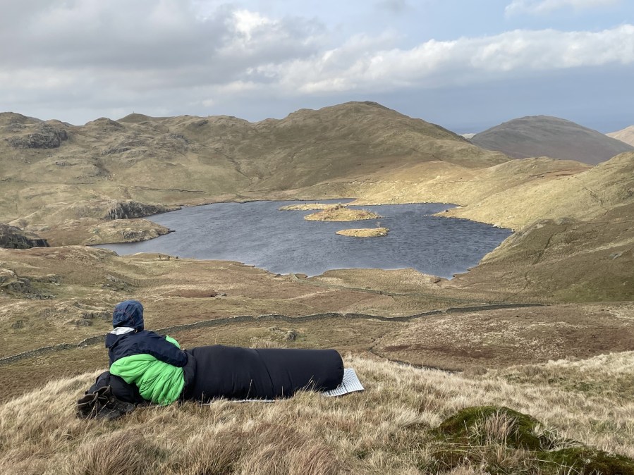 Camper in a green and neon green sleeping bag lying above Angle Tarn lake, Lake District