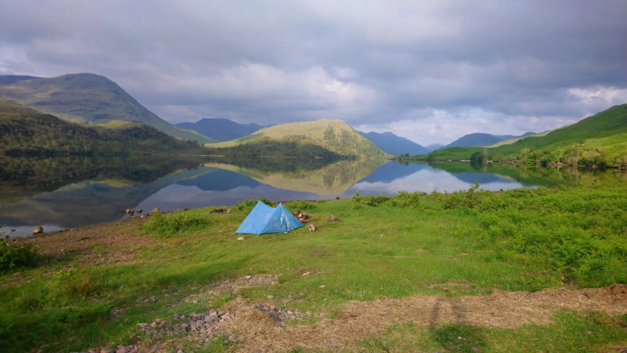 Bright blue tent constructed beside Loch Arkaig with mountains in background