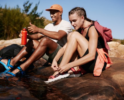 Man and woman in the sun, sat on a brown rock with Lizard sandals on feet dipping in the water