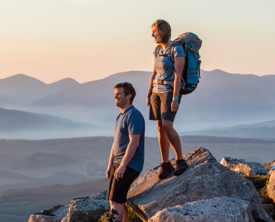 Woman standing on a rock above a man smiling looking into the distance, clouded mountainous background