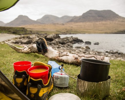 Gas cooker and shoes holding pots of soup outside tent next to lake